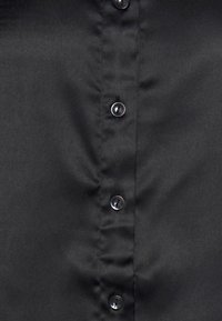 Camicia nera con bottoni a pressione e una texture liscia e opaca. Presenta bottoni trasparenti lungo la parte anteriore, creando un design simmetrico.