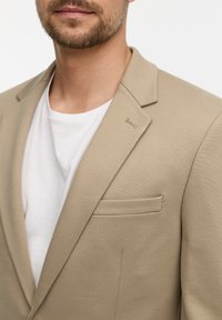 Beige Blazer aus glattem Stoff, mit Spitzrevers, einer einzelnen Brusttasche und einem weißen Rundhals-T-Shirt darunter.
