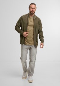 Olivgrön zip-up kofta med ribbade manschetter, lagd över en beige T-shirt. Ljusgrå jeans och beiga sneakers fulländar looken.