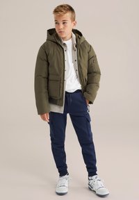 Olivnozeleni pufast plašč s kapuco, sivi notranji sloj, bela srajca in mornarsko modri trenirke. Tenisice z modrimi poudarki dopolnjujejo videz.