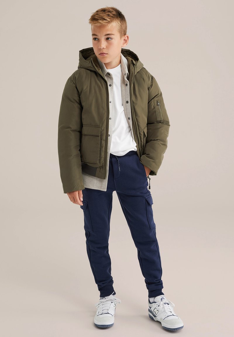 Olivnozeleni pufast plašč s kapuco, sivi notranji sloj, bela srajca in mornarsko modri trenirke. Tenisice z modrimi poudarki dopolnjujejo videz.