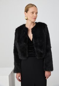 Kvinne som har på seg svart faux fur-jakke, svart kjole og store runde gulløredobber, ser svakt mot siden foran en ensfarget bakgrunn.