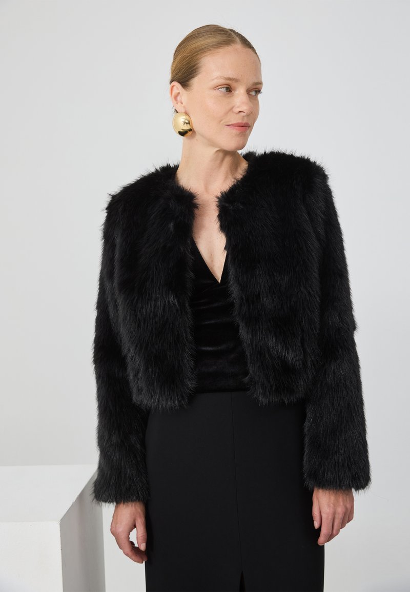 Kvinne som har på seg svart faux fur-jakke, svart kjole og store runde gulløredobber, ser svakt mot siden foran en ensfarget bakgrunn.