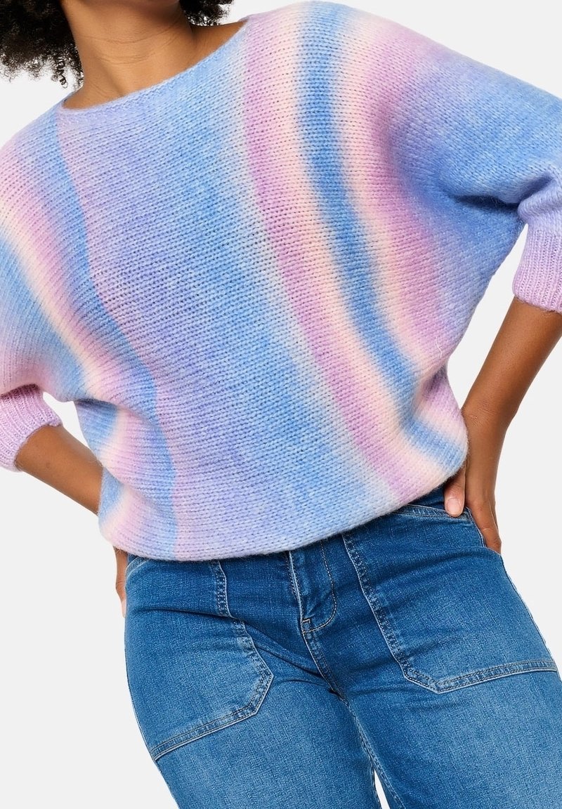Pletený svetr v gradientních odstínech modré, růžové a levandulové. Má volný střih, široký výstřih a tříčtvrteční rukávy, kombinovaný s modrými džíny.