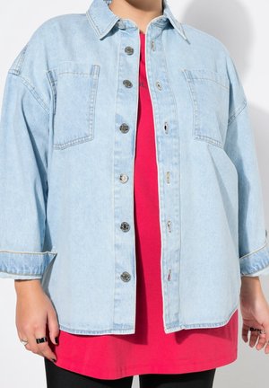 Person iført en lyseblå oversized denimjakke med knapper over en knaldrød skjorte og sorte bukser.