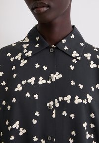 Zwarte blouse met witte vlinderprint, met kraag, korte mouwen en knoopsluiting, met een gladde textuur en subtiele glans.