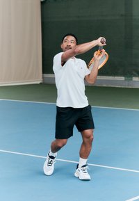Eine Person in einem weißen Poloshirt und schwarzen Shorts schwingt einen Schläger mit einem orangefarbenen Kopf auf einem blauen Tennisplatz. Weiße und marineblaue Sportschuhe.
