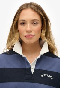 Navy und hellblau gestreiftes Poloshirt mit weißem Kragen. Verfügt über eine Knopfleiste und ein "SUPERDRY"-Logo auf der Brust. Baumwollmaterial.