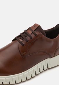 Brown leather shoe featuring a thick white rubber sole, rounded shape, and detailed stitching along the sides. Brown laces complete the design.