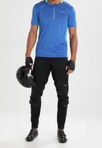 Jersey de ciclismo azul de manga corta con cuello de cremallera y acentos verdes; combinado con pantalones y guantes negros, sosteniendo un casco negro.