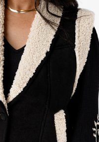 Zwarte vest met crèmekleurige schapenvacht kraag, met een knoopdetail en gestructureerde stof. Close-up toont het gelaagde textielontwerp.