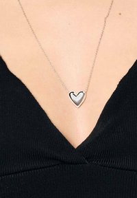 Srebrny wisiorek w kształcie serca na delikatnym łańcuszku, z gładkim wykończeniem i lekkim połyskiem, noszony na tle czarnego materiału.