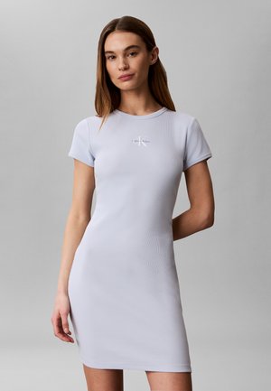 Femme en robe ajustée bleu clair à manches courtes avec logo Calvin Klein sur la poitrine, debout devant un fond gris uni.
