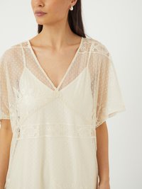 Blouse en dentelle ivoire avec un tissu transparent et texturé, décolleté en V et manches courtes à volants. Présente des motifs floraux et géométriques complexes.