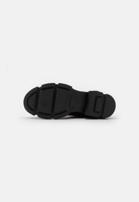 Svart gummisåle med teksturert overflate, som har et segmentert design og forsterket hæl for holdbarhet.