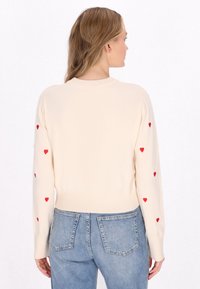 Maglione beige a maglia con maniche lunghe, caratterizzato da ricami a forma di cuore rosso lungo le maniche. Orlo e collo a costine, design corto.