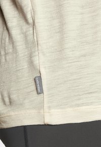 Camicia beige chiaro realizzata in un tessuto morbido e texturizzato con un'etichetta visibile. L'orlo incontra una sezione di tessuto scuro sottostante.