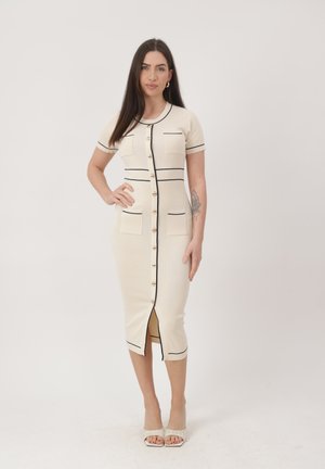 Vrouw met lang donker haar in een beige midi-jurk met knopen, zwarte bies en korte mouwen, staand met één hand op de heup, met open schoenen met hak.