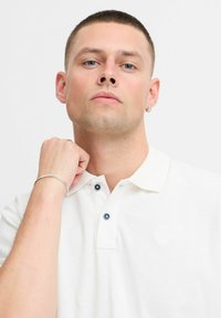 Hvid polo-tee med to marineblå knapper, tekstureret stof og et diskret logo. Modellen har et sølvarmbånd på og poserer med hånden på hagen.