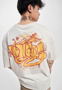 Overdimensioneret cremet t-shirt med fed rød og gul graffiti-stil tekst og et slangedesign på ryggen, med korte ærmer og en afslappet pasform.