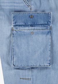 Tissu denim bleu clair avec une grande poche carrée équipée d'un rabat et fermée par des boutons pression métalliques.