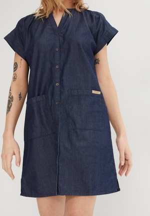 Vrouw draagt een donker denim jurk met korte mouwen en knopen aan de voorkant, voorzien van twee voorzakken en zichtbare getatoeëerde armen.