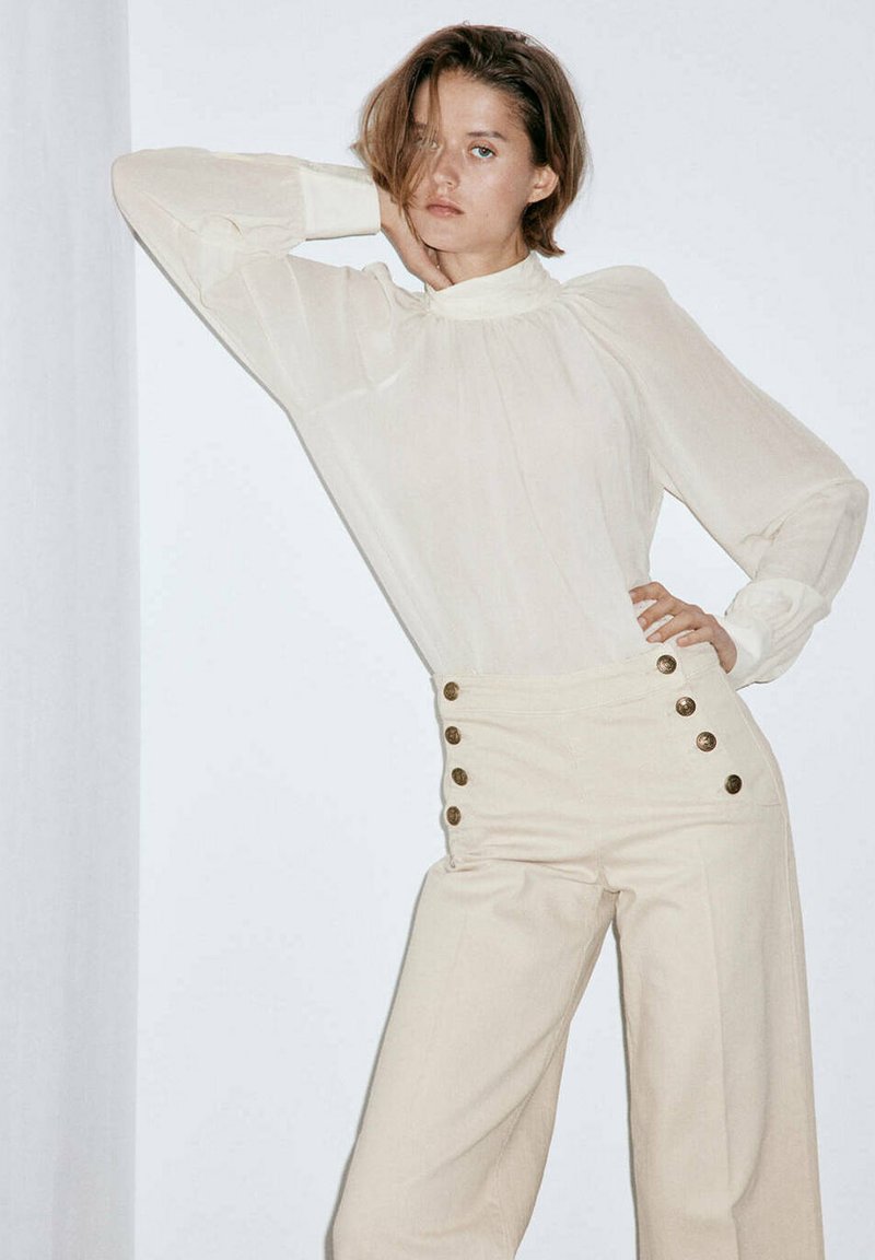 Femme portant un pantalon beige taille haute à six boutons et un chemisier crème transparent à manches longues, posant avec une main sur la hanche et l'autre touchant son cou.