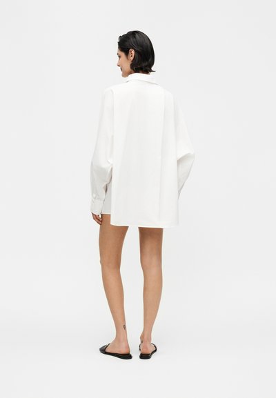 Γυναίκα με κοντά σκούρα μαλλιά που φοράει ένα υπερμεγέθες λευκό μακρυμάνικο πουκάμισο και μαύρα παπούτσια χωρίς κορδόνια, στέκεται γυρισμένη με την πλάτη σε λευκό φόντο.