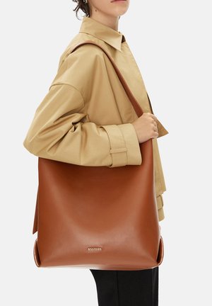 Persona con chaqueta beige sostiene un gran bolso de hombro de cuero marrón con el logo "Gino Rossi" en el frente.