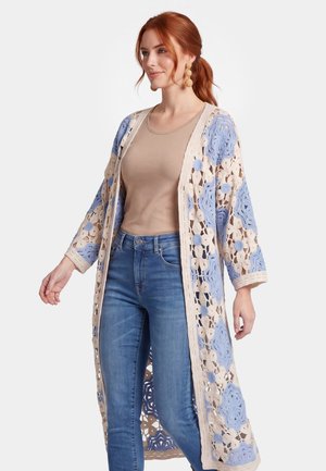 Vrouw met rood haar, die een beige top, blauwe jeans en een lange beige en blauwe gebloemde gehaakte cardigan draagt, staand tegen een effen achtergrond.