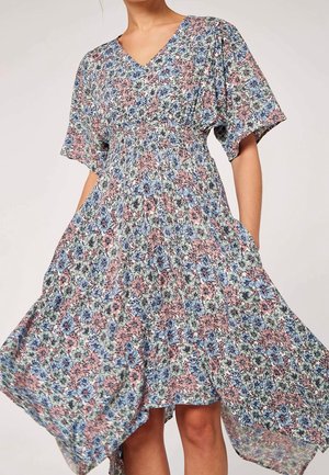 Kvinde iført en knælængde blomsterkjole med korte ærmer, V-hals og samlet talje, holder siderne, mens stoffet flyder udad.