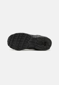 Semelle de chaussure de sport noire avec un motif de semelle texturé, présentant des rainures et des crampons arrondis pour une meilleure adhérence. Logo "New Balance" visible.