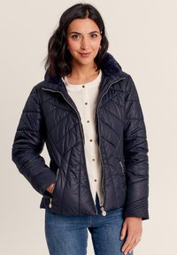 Marineblaue Steppjacke mit Rautensteppung, Kunstpelzkragen, Frontreißverschluss und Seitentaschen. Getragen über einem weißen Oberteil und blauen Jeans.