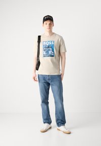 Tricou bej cu un imprimeu grafic albastru care reprezintă un vehicul, purtat împreună cu blugi albaștri și adidași albi. Caciulă neagră cu logo.