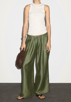 Persoon draagt een mouwloos wit topje, losse olijfgroene wijde broek, houdt een bruine met kralen versierde tas vast en draagt zwarte sandalen.