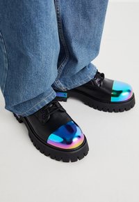 Sapatos de couro pretos com bicos redondos iridescentes, solas chunky pretas e atacadores pretos, combinados com calças de denim azuis.