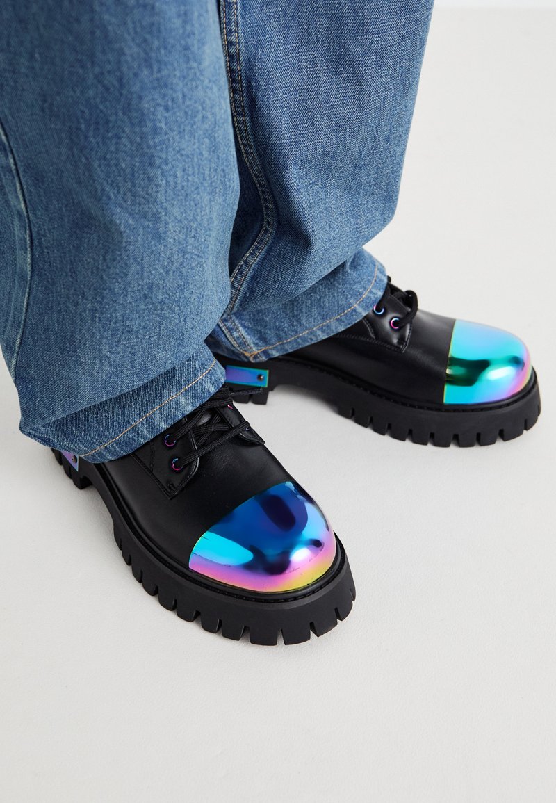 Sapatos de couro pretos com bicos redondos iridescentes, solas chunky pretas e atacadores pretos, combinados com calças de denim azuis.