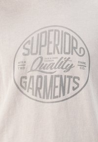 Lys grå t-skjorte med et stort ovalt grafikk i mørkere grå tekst som leser "Superior Quality Garments" med merkeinformasjon inkludert.