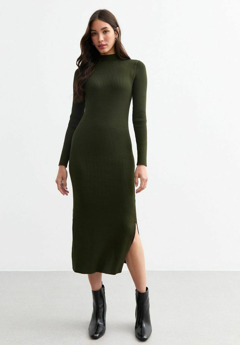 Robe en tricot côtelé vert olive avec des manches longues, un col montant, une fente latérale et une silhouette ajustée, associée à des bottines noires.