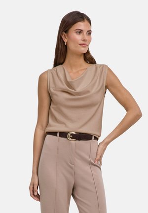 Femme portant un haut beige sans manches à encolure drapée et un pantalon beige taille haute, posant avec une main sur la hanche sur fond blanc.