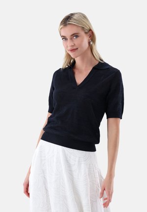 Blonde Frau trägt ein schwarzes V-Ausschnitt-Oberteil mit kurzen Ärmeln und einen weißen Rock mit Muster, stehend vor einem einfarbigen hellen Hintergrund.