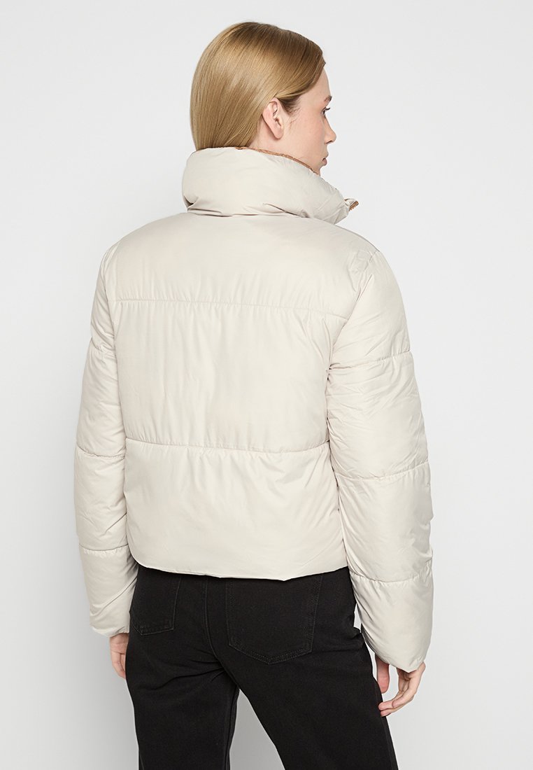 Chaqueta acolchada beige claro con cuello alto, diseño acolchado y corte cropped. Presenta una textura suave y un estilo minimalista.