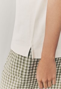 T-shirt bianca in cotone con spacchi laterali, abbinata a una gonna a quadri verde e bianca. Il tessuto sembra morbido e leggero.
