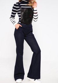 Zwarte leren tuinbroek met verstelbare bandjes, gecombineerd met een marineblauwe en witte gestreepte coltrui. Uitlopende donkere spijkerbroeken, witte sneakers.