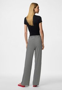 Pitkät blondit hiukset omaava nainen, jolla on musta istuva yläosa, mustavalkoiset vaakaraitaiset leveälahkeiset housut ja punaiset sandaalit, seisomassa selin.