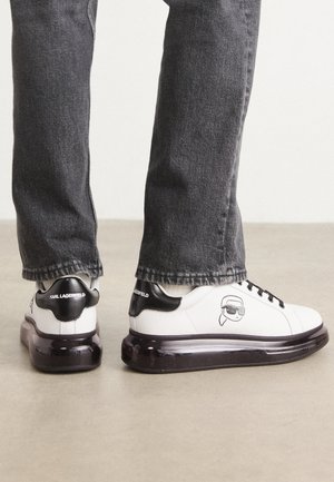 Zapatillas blancas con un acento negro en el talón y logo, que presentan un diseño gráfico en el lateral y una suela gruesa y transparente.