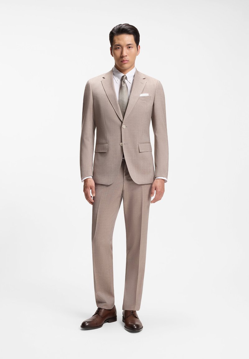 Homme debout portant un costume beige avec une chemise blanche, une cravate gris clair, une pochette blanche et des chaussures marron sur un fond blanc uni.