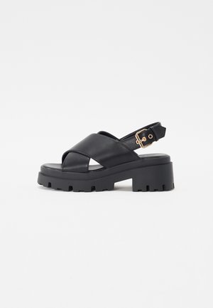 Sandale din piele neagră cu un design cu curea încrucișată, talpă platformă neagră masivă și curea ajustabilă la gleznă cu accente de hardă de aur.