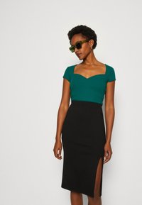 Teal geribbelde crop top met een vierkante halslijn, gecombineerd met een zwarte rok tot de knie met een zijsplit. Model draagt groene zonnebril.