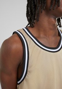 Beige netting tank topp med svart og hvit stripet kant. Har rund hals og er uten ermer, og viser en pustende tekstur.
